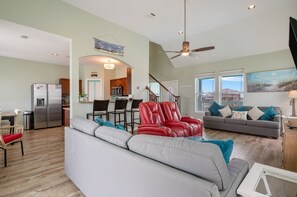 Un espacio habitable espacioso con un sofá gris, un sillón reclinable rojo y un sofá gris con cojines verde azulado. Se ve una cocina con refrigerador y microondas al fondo.
