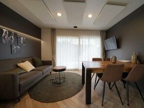 A modern living room with a sofa, a round coffee table, a dining table with chairs, a TV mounted on the wall, and a large window with curtains. - Lieteberg Cottage 4 (Zutendaal)