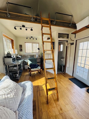 A room with a wooden ladder, a bed, a kitchenette, and a door leading outside.