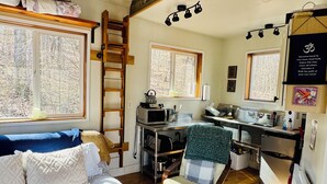 A cozy cabin interior with a loft bed, a dining area, a kitchen, and a seating area with a view of the forest.