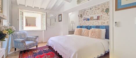 A bedroom with a bed, a chair, a window, a mirror, and a wall with a shelf.