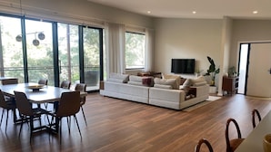 A modern living room with a dining area, large windows, and a flat-screen TV. - Escape to Nature in Our Warm & Welcoming Family Home (Warrandyte)
