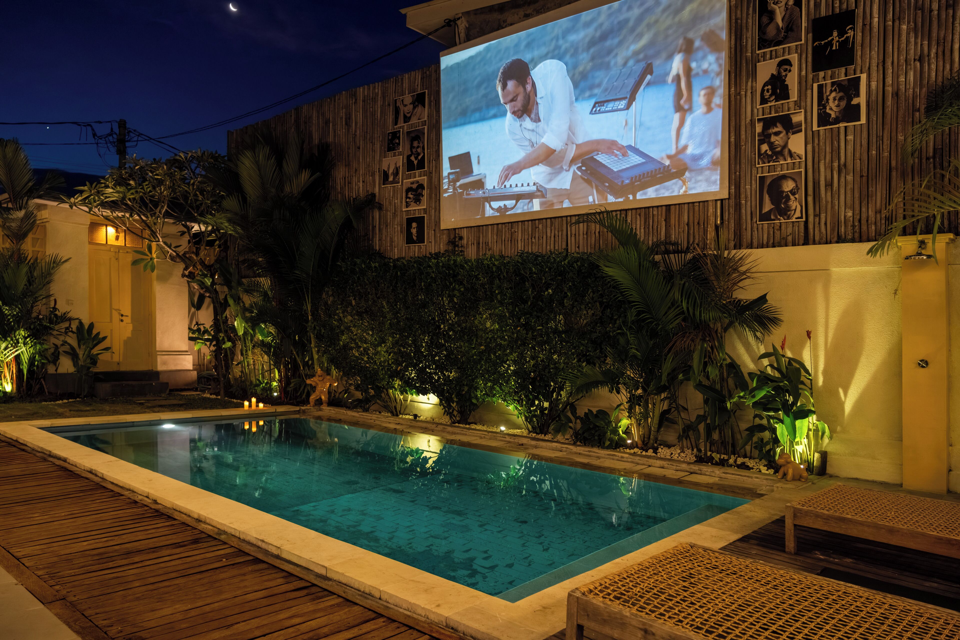 A pool area with a wooden deck, lounge chairs, and a large screen displaying a movie.
