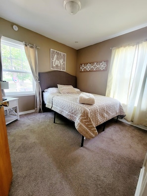 A bedroom with a bed, a bedside table, a wall decoration, and a window with curtains. - Canyon Lake GETAWAY 2 bedrooms-1-KING & 1 QUEEN (Rapid City)