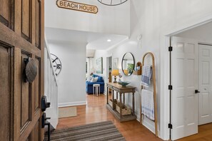 Ein Flur mit einer Holztür, einem Schild mit der Aufschrift 'BEACH HOUSE', einem blauen Sofa, einem kleinen Tisch mit einer Lampe und einem Spiegel an der Wand.