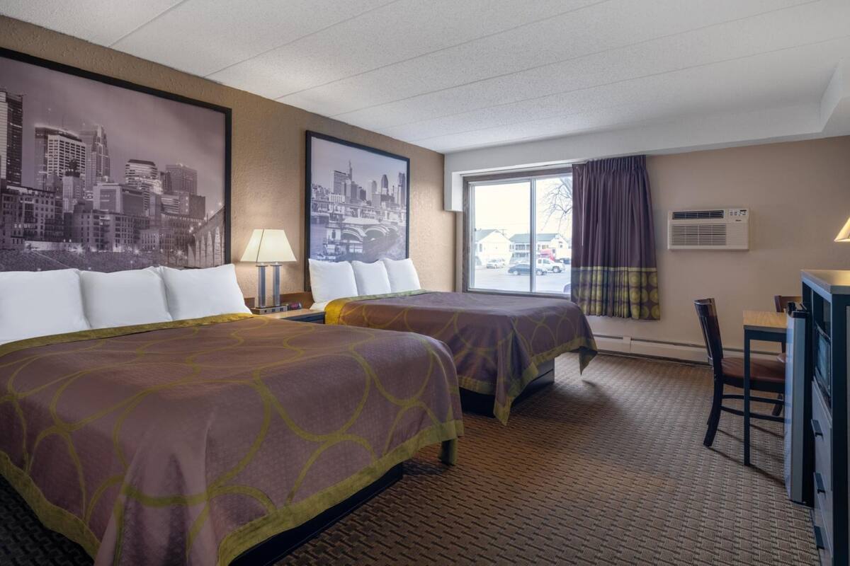 A hotel room with two beds, a desk, a chair, a window with curtains, and a cityscape artwork on the wall.