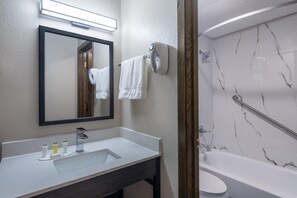 A bathroom with a sink, mirror, towel rack, and a bathtub with marble walls. - Coratel Inn & Suites by Jasper Owatonna (Owatonna)