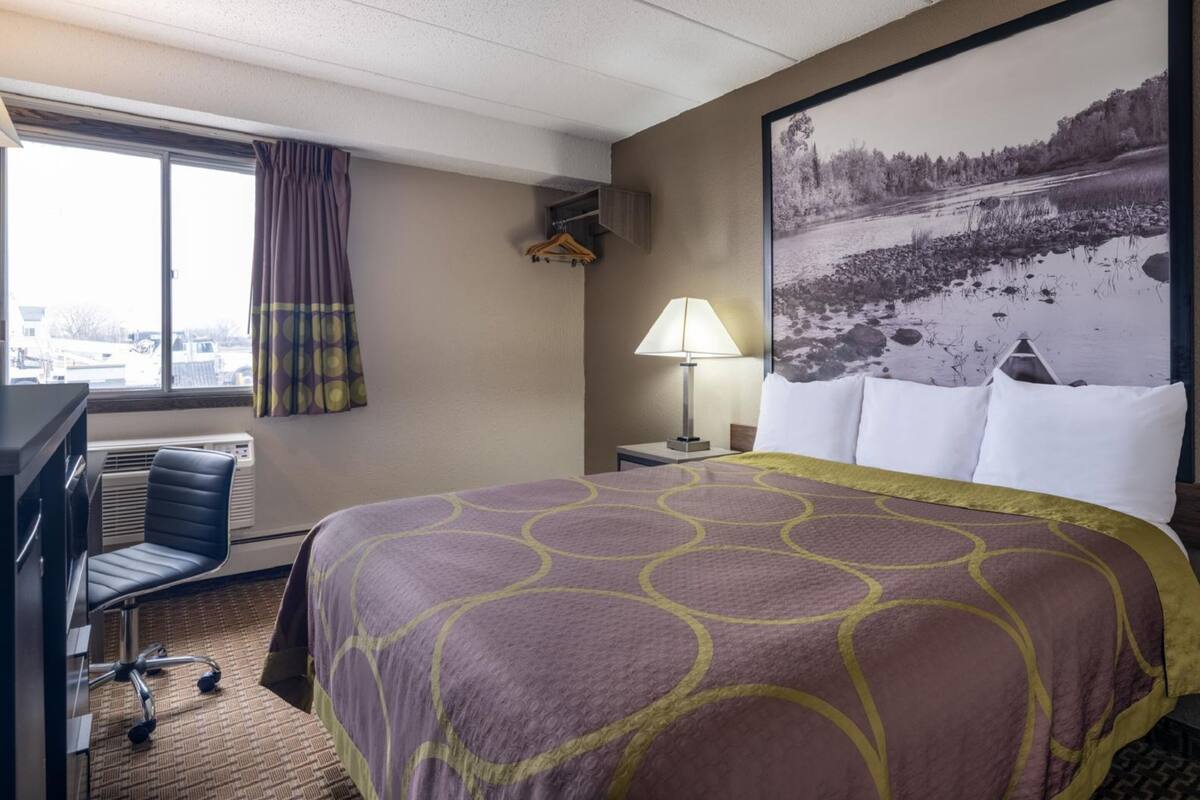 A hotel room with a large bed, a desk with a chair, a window with curtains, a lamp, and a wall-mounted coat rack.
