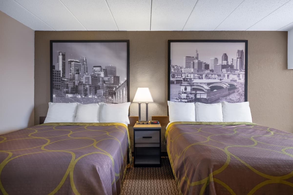 A hotel room with two beds, a nightstand with a lamp, and two cityscape posters on the wall.
