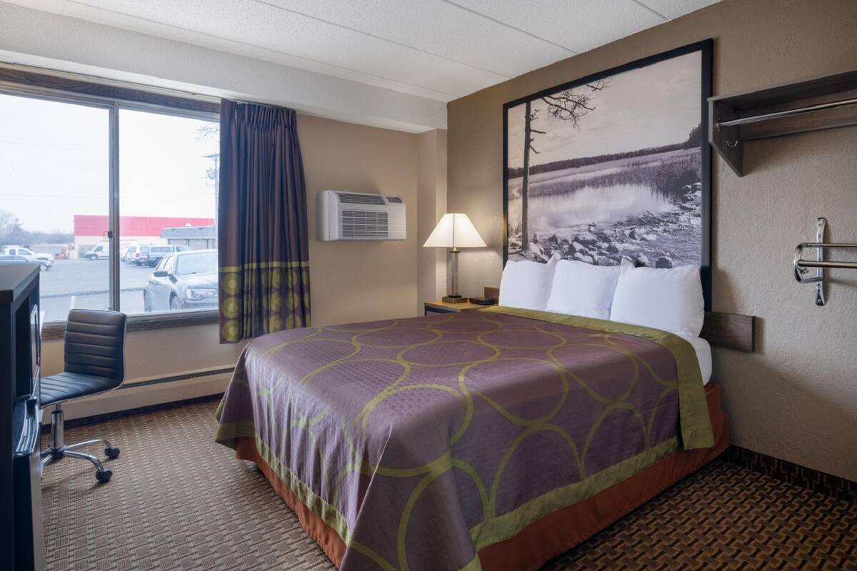A hotel room with a bed, a desk, a chair, a window with curtains, an air conditioner, a lamp, and a painting on the wall.