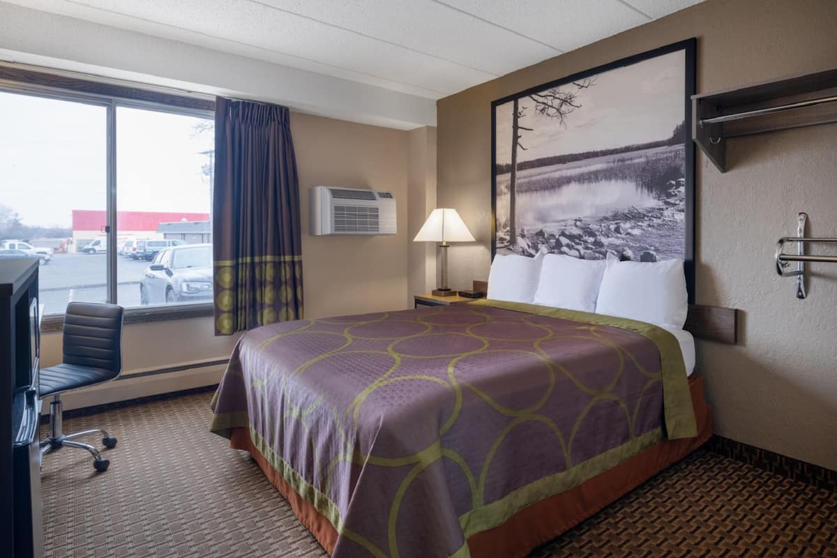 Habitación de hotel con cama, escritorio, silla, ventana con cortinas, aire acondicionado, lámpara y un cuadro en la pared.