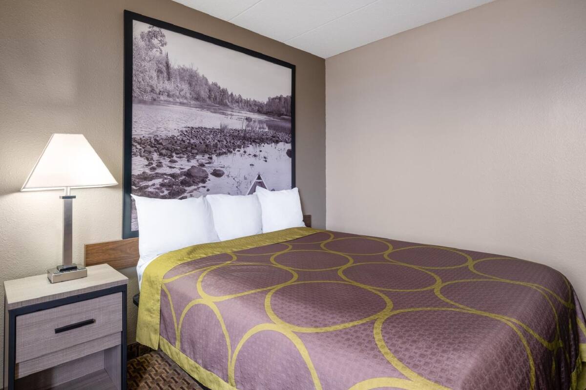 A hotel room with a bed, a nightstand with a lamp, and a large framed picture of a river landscape.