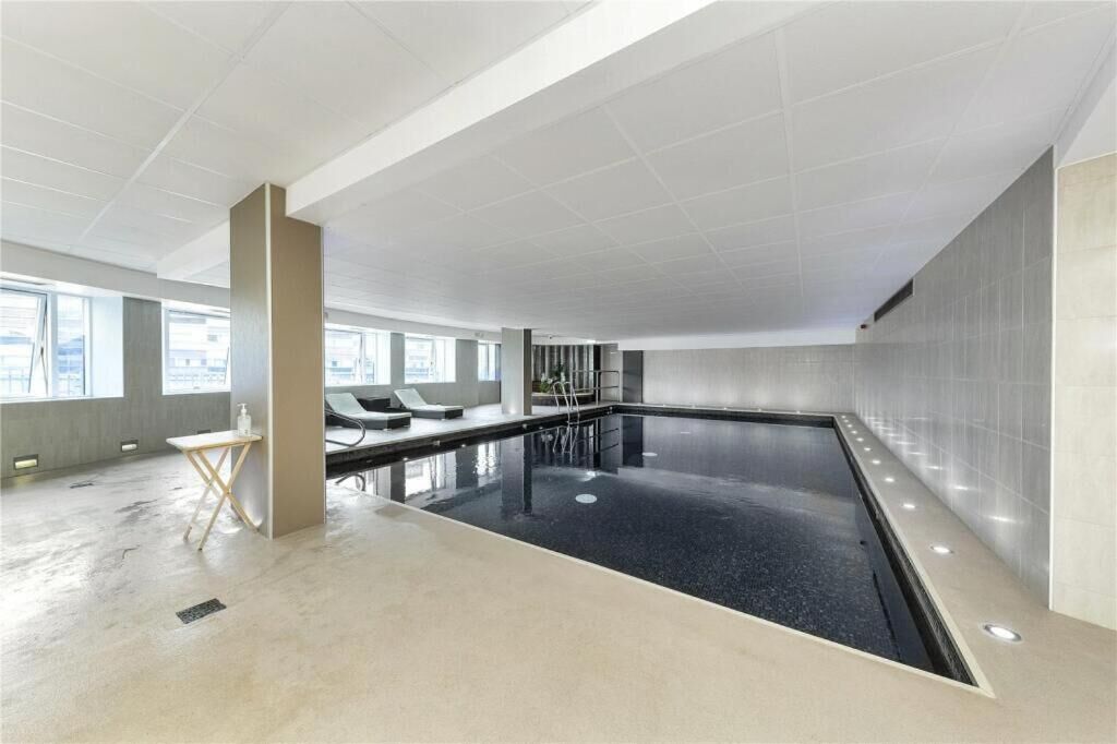 A modern indoor swimming pool with a tiled floor, a lounge area with seating, and large windows.