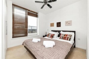 A bedroom with a bed, a ceiling fan, a window with blinds, and wall art. - The Luxurious Penthouse (Cambridge)