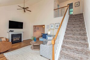 A living room with a fireplace, a television, a staircase, and a ceiling fan. - Ocean Pines Home with Sunroom and Beach Pass (Ocean Pines)