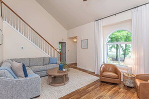 A living room with a staircase, a large window with curtains, a sofa, a coffee table, and a lamp. - Ocean Pines Home with Sunroom and Beach Pass (Ocean Pines)