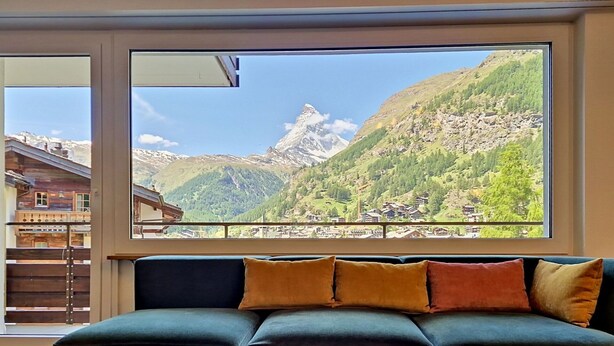 A room with a large window overlooking a mountainous landscape, a dark green sofa with cushions, and a small white table with a book and two circular objects. - Lovely apartment for 4 people with WIFI, TV and balcony (Zermatt)
