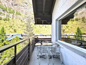 Ein Balkon mit Tisch und Stühlen, mit Blick auf eine gebirgige Landschaft und Gebäude in der Ferne.