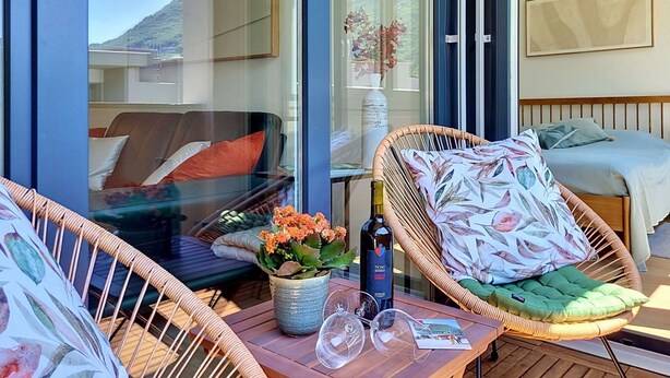 Een balkon met rieten meubilair, een tafel met een fles en glazen, en een bed met een bloemenkussen.