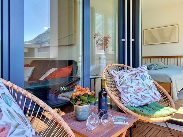 A balcony with wicker furniture, a table with a bottle and glasses, and a bed with a floral pillow.