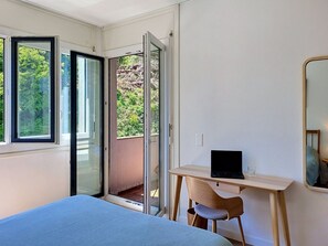 Ein Schlafzimmer mit einem Bett, einem Schreibtisch mit Laptop und einer Glasschiebetür, die zu einem Balkon führt.