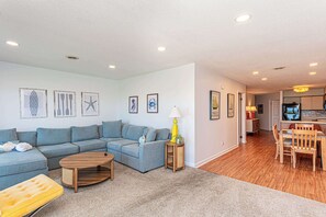 A living room with a sofa, coffee table, and lamp; a dining area with chairs and a table; and a kitchen with a refrigerator and counter. - Bayfront 2BR Condo with Water Views (Ocean City)
