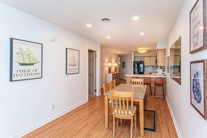 A dining area with wooden chairs and a table, a kitchen with a refrigerator and counter, and framed artwork on the walls. - Bayfront 2BR Condo with Water Views (Ocean City)