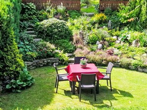 Un patio jardín con mesa y sillas, rodeado de abundante follaje y un muro de piedra.