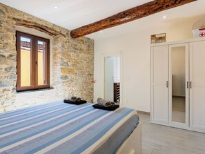 Ein Schlafzimmer mit Steinsandsteinwand, Holzbalkendecke, einem Bett mit gestreifter Bettwäsche, einem Kleiderschrank und einem Fenster mit Blick.