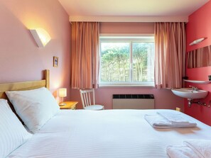Ein Hotelzimmer mit Bett, Nachttisch mit Lampe, Stuhl, Fenster mit Vorhängen, Waschbecken und Spiegel.