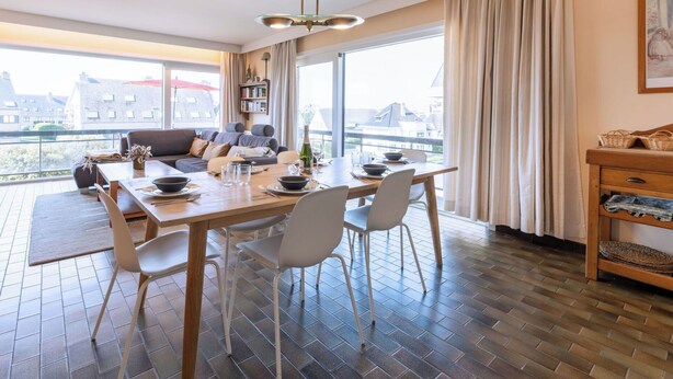 Een moderne woonkamer met een eethoek, grote ramen en uitzicht op gebouwen.