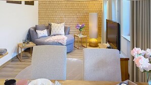 A modern living room with a sofa, two armchairs, a TV, a lamp, and a brick wall. - Cosy apartment for 2 guests with WIFI, TV and patio (Loch)