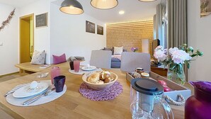 A modern living room with a wooden dining table, a sofa, and a TV. There are three pendant lights, a brick wall, and a window with curtains. - Cosy apartment for 2 guests with WIFI, TV and patio (Loch)