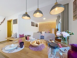 Ein modernes Wohnzimmer mit einem Holzesstisch, einer Couch und einem Fernseher. Drei Pendelleuchten, eine Ziegelwand und ein Fenster mit Vorhängen sind zu sehen.