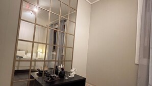 Ein Zimmer mit einem großen Spiegel, einem dunklen Konsolentisch, einem Bett mit weißem Kopfteil und einem Fenster mit Vorhang.