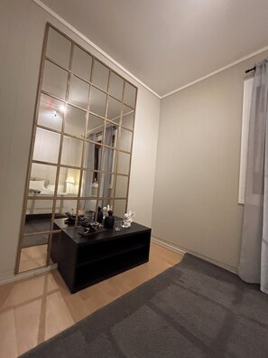 A room with a large mirror, a dark console table, a bed with a white headboard, and a window with a curtain.