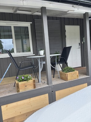 A covered outdoor seating area with a table and chairs, wooden planters with greenery, and a door.