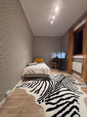 Une chambre avec un papier peint à motifs, un tapis à imprimé zébré, un lit avec des coussins, un bureau avec une chaise et une fenêtre avec des rideaux.