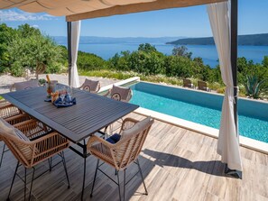 Ein Außenpoolbereich mit Tisch und Stühlen, Blick auf das Meer und einer Pergola.