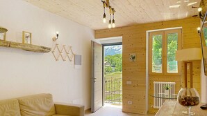 Ein Zimmer mit Holzboden, einem beigefarbenen Sofa, einem Glastisch mit einer Vase und einer Tür, die auf einen Balkon mit Blick auf Grünflächen und Berge führt.