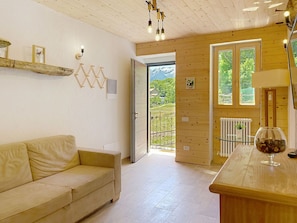 Ein Zimmer mit Holzboden, einem beigefarbenen Sofa, einem Glastisch mit einer Vase und einer Tür, die auf einen Balkon mit Blick auf Grünflächen und Berge führt.