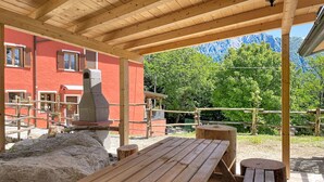 Eine hölzerne Außenbereich-Sitzgelegenheit mit Bänken und einem Tisch, ein Steinkamin und ein rotes Gebäude im Hintergrund.