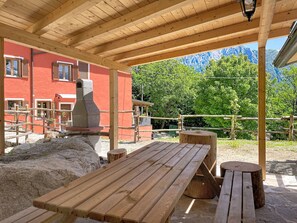Eine hölzerne Außenbereich-Sitzgelegenheit mit Bänken und einem Tisch, ein Steinkamin und ein rotes Gebäude im Hintergrund.