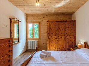 Quarto com teto de madeira, cama com roupa de cama branca, cômoda de madeira, radiador e janela com vista para vegetação.
