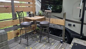 A wooden deck with two metal chairs and a table, a small potted plant, and a window with a view of greenery.