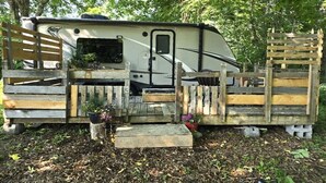 A recreational vehicle with a ramp leading to a wooden deck, surrounded by trees.