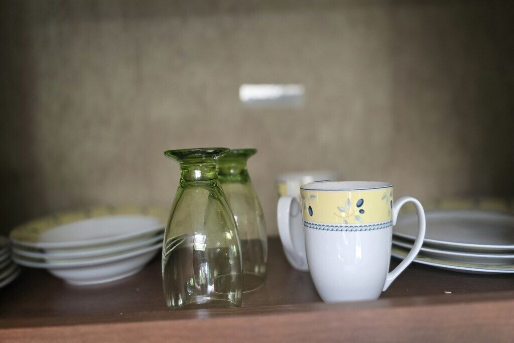 Ein Regal mit gestapelten Tellern, einer klaren Glasgießkanne und einer weißen Tasse mit blauem Muster.