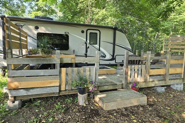 A recreational vehicle (RV) parked in a wooded area with a wooden fence and steps leading to the entrance.