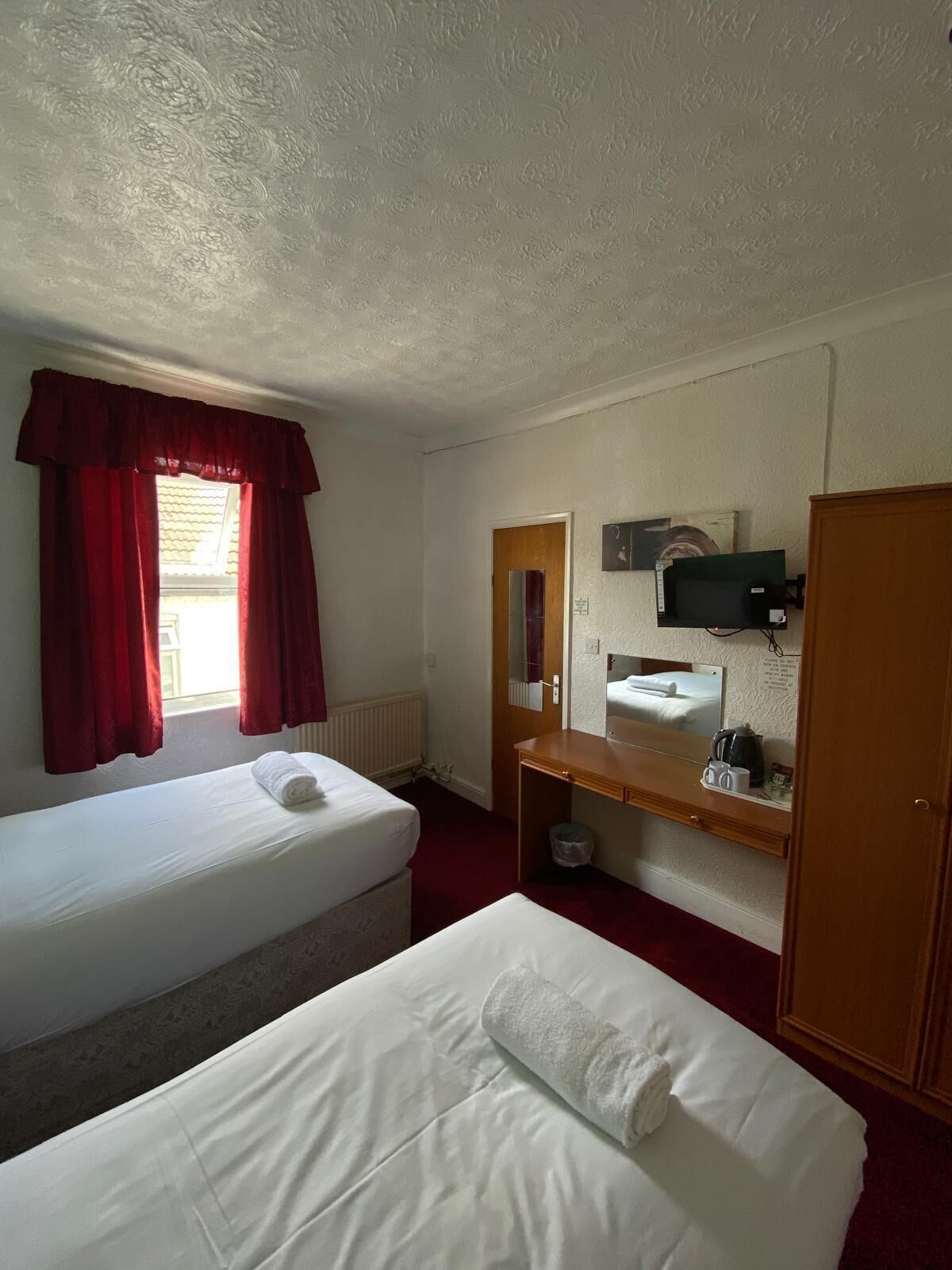 A hotel room with two single beds, a wooden dresser, a mirror, a microwave, and a window with red curtains.