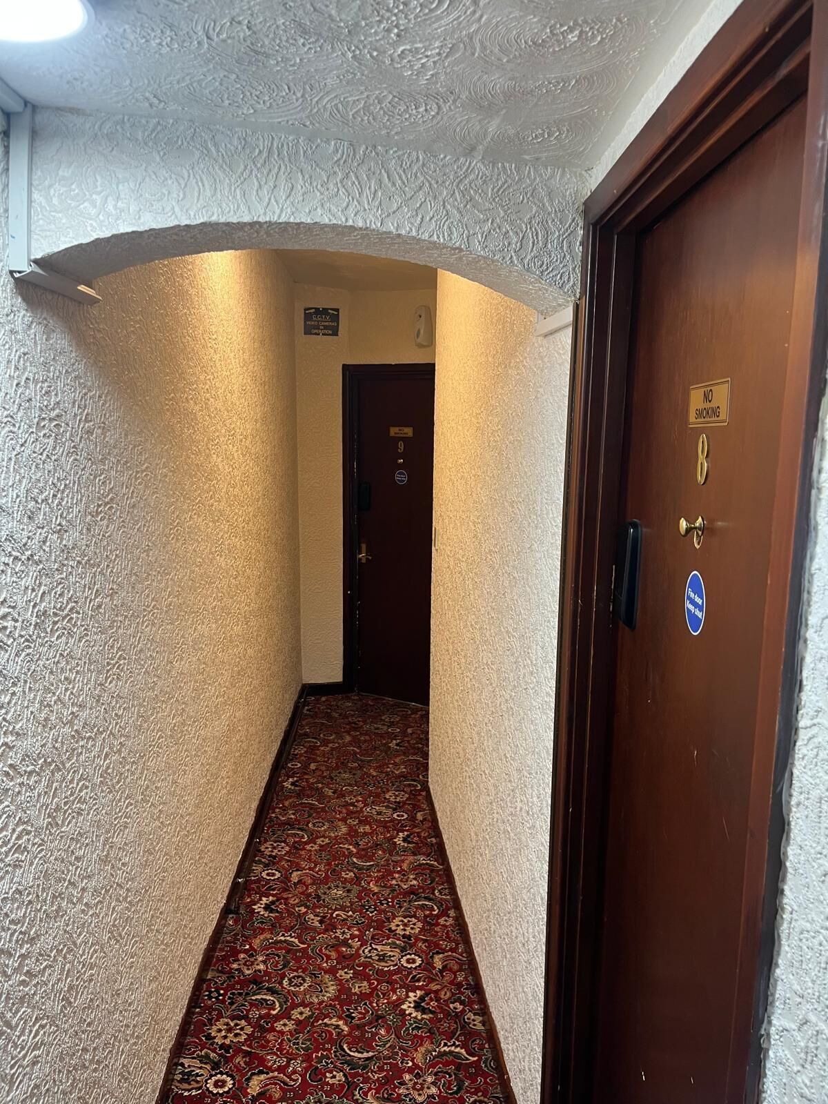 Un couloir aux murs texturés, agrémenté d’une moquette à motifs et doté d’une porte en bois sur laquelle est apposé un panneau « Interdiction de fumer ».